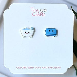 May include: A pair of Tiny Cuts Crafts earrings on a white card. One earring is a white bathtub with blue accents, the other is a blue toaster. The card has the text "Tiny cuts Crafts" and "CREATED WITH LOVE AND PRECISION".