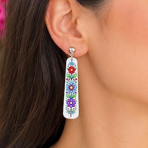 May include: A white, elongated, rectangular drop earring featuring a colorful floral pattern with red, blue, purple, and green petals, accented by small black line details and dots, suspended from a small silver-toned metal ball stud post.