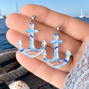 May include: A pair of anchor-shaped earrings with a nautical theme. The earrings feature a blue, white, and tan striped design with a silver post. The earrings are held in a hand, with a blurred background of the ocean and sailboats.