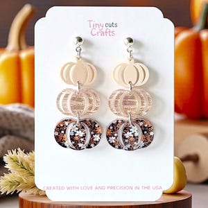 May include: A pair of dangle earrings featuring a three-tiered pumpkin design. The top two tiers are a solid cream color, while the bottom tier showcases a floral pattern with white flowers on a dark background. The earrings are displayed on a white card with the text "Tiny Cuts Crafts".