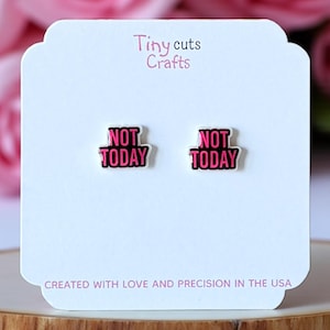 May include: A pair of black and pink "NOT TODAY" earrings on a white card. The card has the text "Tiny cuts Crafts" at the top and "CREATED WITH LOVE AND PRECISION IN THE USA" at the bottom. The earrings are displayed on a wooden surface.