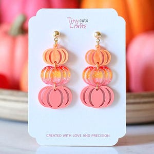 May include: A pair of layered pumpkin earrings in shades of orange and pink, with gold-colored studs. The earrings are displayed on a white card with the text "Tiny cuts Crafts" and "CREATED WITH LOVE AND PRECISION". The background features blurred pumpkins.