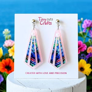 May include: A pair of pink, geometric earrings with a silver stud. The earrings feature a colorful leaf design in blue, green, and white. The text "Tiny cuts Crafts" and "CREATED WITH LOVE AND PRECISION" are visible.