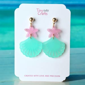 May include: A pair of earrings featuring a pink starfish and a turquoise seashell design. The earrings are hanging from gold-colored posts. The card behind the earrings has the text "Tiny cuts Crafts" and "CREATED WITH LOVE AND PRECISION."