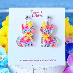 May include: A pair of cat-shaped earrings with a floral pattern in pink, blue, and yellow. Each earring has a yellow bow and a small yellow flower on top. The earrings are displayed on a white card with the text "Tiny cuts Crafts" and "CREATED WITH LOVE AND PRECISION".