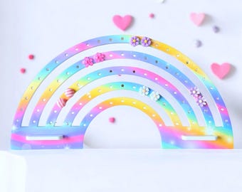 May include: A rainbow-shaped earring holder with multiple rows of holes for displaying jewelry. The holder is painted in a gradient of colors, including pink, yellow, blue, and purple. Small flower-shaped earrings are displayed on the holder.