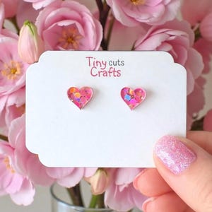 May include: A pair of heart-shaped stud earrings with pink glitter, displayed on a white card with the text "Tiny cuts Crafts". The earrings are held in front of a backdrop of pink roses.