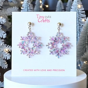 May include: A pair of snowflake-shaped earrings with a silver post and a dangling design. The earrings are made of clear acrylic with iridescent glitter, displaying shades of pink, purple, and silver. The text "Tiny cuts Crafts" and "CREATED WITH LOVE AND PRECISION" are visible.
