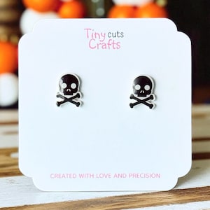 May include: A pair of black and white skull and crossbones earrings on a white card. The card has the text "Tiny cuts Crafts" and "CREATED WITH LOVE AND PRECISION". The earrings are a spooky accessory.