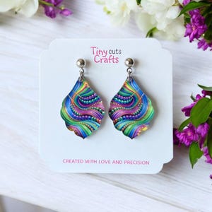 May include: A pair of colorful dangle earrings with a silver hook. The earrings feature a teardrop-shaped design with a swirling pattern of green, blue, purple, and pink. The text "Tiny cuts Crafts" is printed on the white card behind the earrings. The card also says "Created with love and precision."