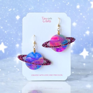 May include: A pair of gold hoop earrings with a pink and blue galaxy-themed planet charm. The planet has a ring around it and is covered in glitter. The earrings are on a white card with the text "Tiny cuts Crafts" and "Created with love and precision".