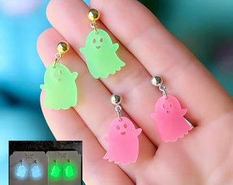 May include: A pair of green and a pair of pink ghost-shaped earrings. The earrings have a gold or silver-colored post and a small, smiling ghost charm. The earrings are displayed on a hand.