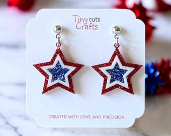 Puede incluir: Pendientes en forma de estrella con bordes rojos brillantes, interior blanco y centros azules brillantes. Los pendientes se muestran en una tarjeta blanca con el texto "Tiny cuts Crafts" y "CREATED WITH LOVE AND PRECISION".
