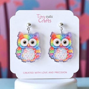 May include: A pair of colorful owl earrings. Each earring features a rainbow-colored owl design with large eyes and a patchwork pattern on the chest. The earrings are displayed on a white card with the text "Tiny cuts Crafts" and "CREATED WITH LOVE AND PRECISION."