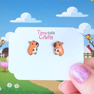 May include: A pair of orange capybara stud earrings on a white card with the text "Tiny cuts Crafts". The earrings are held in front of a cartoon farm scene with a blue sky and white clouds.