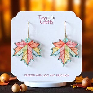 May include: A pair of maple leaf earrings with a colorful patchwork design. The leaves are in shades of red, orange, yellow, and green, with a light blue outline. The earrings hang from gold-colored hoops. The text "Tiny cuts Crafts" is visible.
