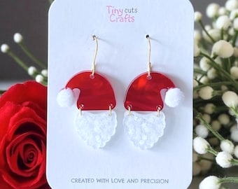 May include: Red and white Santa hat earrings with gold-colored hooks. The earrings are on a white card with the text "Tiny cuts Crafts" and "CREATED WITH LOVE AND PRECISION".