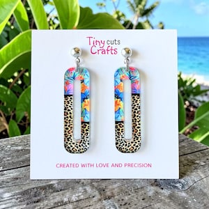 May include: A pair of acrylic earrings with a tropical floral and leopard print design. The earrings are rectangular with a cutout center. The top section features a colorful floral pattern, while the bottom has a leopard print. The text "Tiny cuts Crafts" and "CREATED WITH LOVE AND PRECISION" are visible.