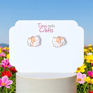 May include: A pair of cat-shaped earrings on a white display card. The earrings feature a sleeping cat design with orange and white coloring. The card has the text "Tiny cuts Crafts" in pink. The background is a field of colorful flowers.