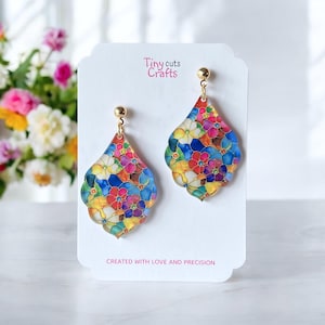 May include: A pair of dangle earrings with a teardrop-shaped design. The earrings are made of clear acrylic with a colorful floral pattern. The earrings have gold-toned hooks.