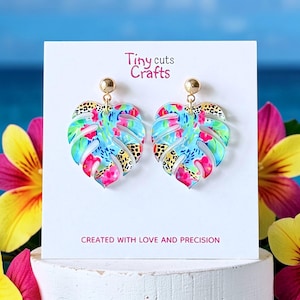 May include: A pair of leaf-shaped earrings with a colorful floral and leopard print design. The earrings are made of a clear material with gold-colored posts. The text "Tiny cuts Crafts" and "CREATED WITH LOVE AND PRECISION" are visible on the white card.