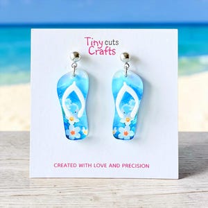 May include: A pair of dangle earrings shaped like blue flip-flops with white thong straps. The earrings feature a sky and ocean design with white flowers. The text "Tiny cuts Crafts" and "CREATED WITH LOVE AND PRECISION" are visible.