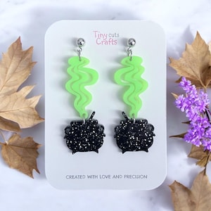 May include: A pair of dangle earrings featuring a black cauldron with white speckles, topped with a neon green smoke design. The earrings are attached to silver-colored posts. The text "Tiny cuts Crafts" and "CREATED WITH LOVE AND PRECISION" are visible.