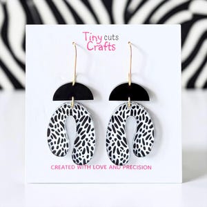 May include: A pair of modern earrings with a black and white spotted design. The earrings have a black semi-circle top and a U-shaped bottom with a black and white pattern. The text "Tiny cuts Crafts" and "CREATED WITH LOVE AND PRECISION" are visible.