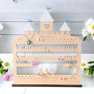 May include: Wooden jewelry organizer in the shape of a castle. The organizer has three white turrets with blue flags, a heart-shaped cutout, and the name "Amelia" engraved. It features multiple slots and bars for hanging jewelry.