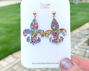 May include: A pair of gold fleur-de-lis earrings with a colorful confetti design. The earrings are on a white card with the text "Tiny cuts Crafts" and "Etsy.com/shop/TinyCutsCrafts".