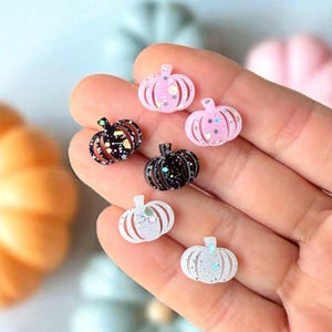 May include: A collection of small, decorative pumpkin-shaped embellishments in various colors. The pumpkins are in shades of pink, black, and white, with glitter accents. These miniature pumpkins are ideal for crafting and seasonal decor.