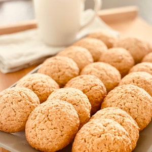Peut inclure: Un plateau rempli de biscuits dorés et fraîchement cuits. Les biscuits ont une surface texturée et sont disposés en rangées. Une tasse blanche et un tissu plié sont en arrière-plan, suggérant un cadre confortable. Les biscuits semblent être à l'avoine ou similaires.