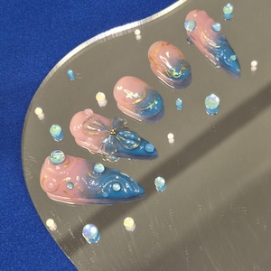 Puede incluir: Cinco uñas postizas largas y puntiagudas con un diseño degradado de rosa y azul. Las uñas están decoradas con purpurina dorada, pedrería y un pequeño dije de mariposa.