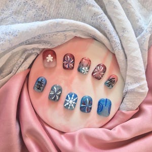 Puede incluir: Un conjunto de uñas artificiales pintadas dispuestas sobre una superficie circular rosa. Las uñas presentan varios diseños, incluyendo patrones florales, copos de nieve y caracteres japoneses, en tonos de rojo, azul, blanco y gris. El fondo incluye tela rosa y blanca.