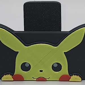 May include: A black phone stand with a yellow Pikachu character peeking out from behind it.