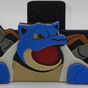 May include: A blue and brown 3D printed figurine of a Pokemon character with a large mouth and two cannons on the sides. The character is on a black background.