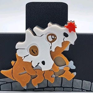 May include: A layered, 3D-printed Cubone and Marowak design. The figures are primarily orange, white, and brown, with a black background and a stone-like base. A small red accent is on top of the skull.