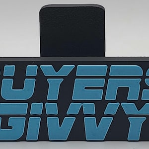 May include: A black rubber bag clip with the words "BUYERS GIVVY" in light blue, bold, block letters. The clip has a rectangular base and a raised, textured handle.