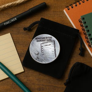May include: A silver-colored coin with the text "STARTING IS THE HARDEST PART" and a to-do list graphic. The coin is in a black drawstring bag, with a black pen and notepads in the background.