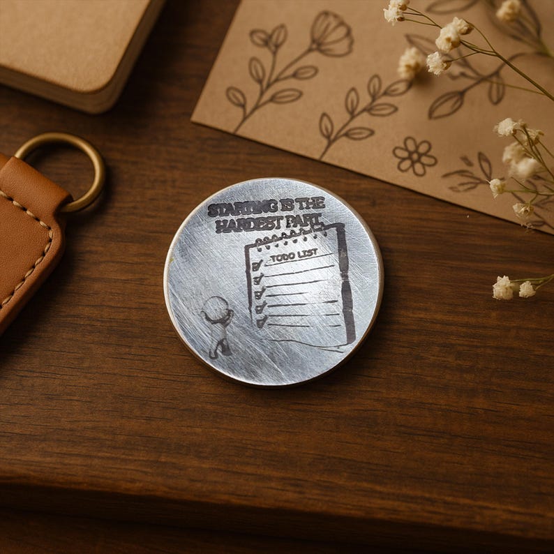 May include: A round, silver-colored metal coin with the text "STARTING IS THE HARDEST PART" and a to-do list graphic. The coin is on a wooden surface, with a leather keychain and a small notebook visible in the background.