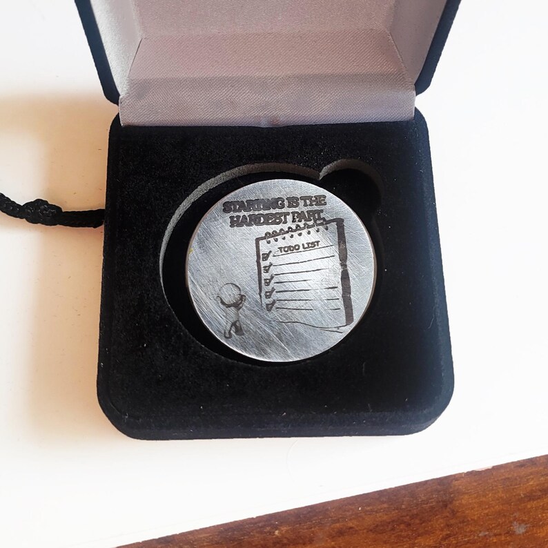 May include: A silver-colored round pendant with the text "STARTING IS THE HARDEST PART" and a to-do list design. The pendant is displayed in a black velvet box with a black cord. The pendant is a motivational gift.