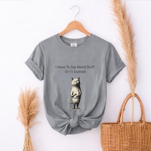 May include: Gray t-shirt with rolled sleeves and a knotted hem. The shirt features the text "I Have To Say Weird Stuff Or I'll Explode" above a cartoon cat holding a book. The cat has large eyes and a serious expression.