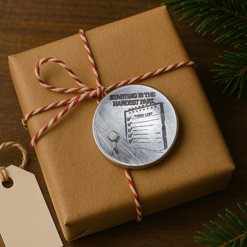 May include: A wrapped gift in brown paper, tied with red and cream twine. A silver-colored circular tag is attached, engraved with the words "Starting is the Hardest Part" and a to-do list graphic. A blank tag hangs nearby.