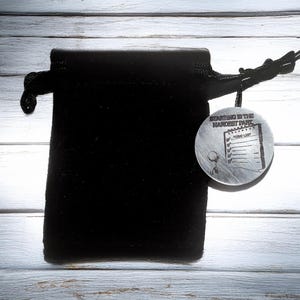May include: A black velvet drawstring bag with a silver-colored round pendant. The pendant has the text "Starting is the Hardest Part" and a to-do list design. The bag is on a light wood-grain surface.