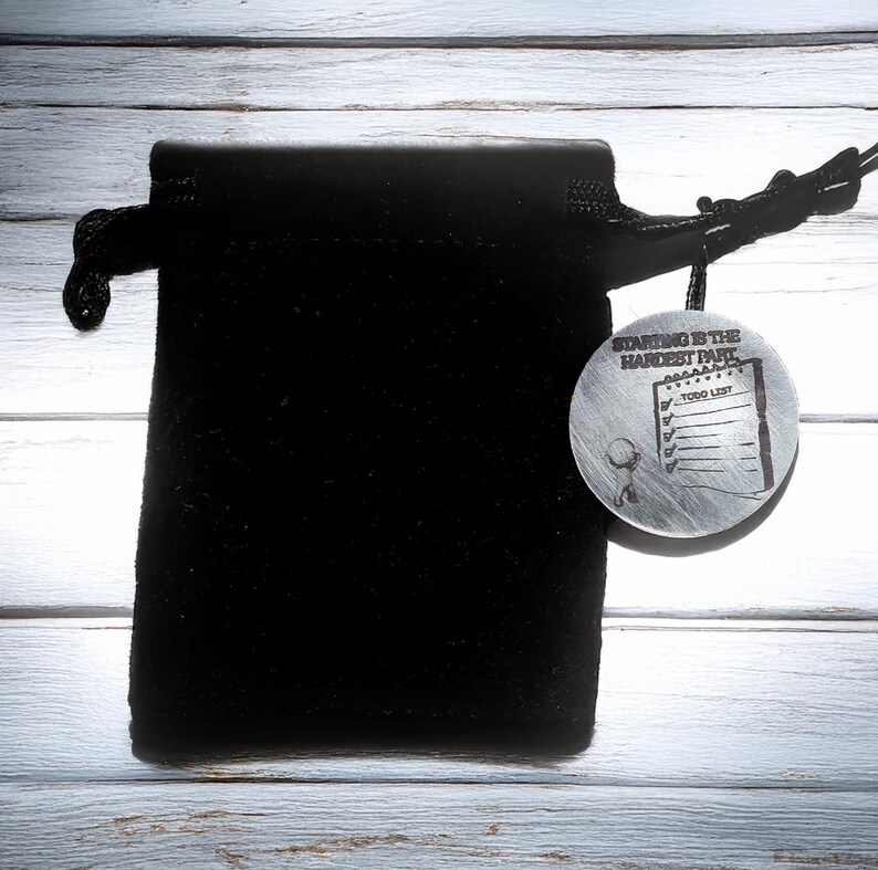 May include: A black velvet drawstring bag with a silver-colored round pendant. The pendant has the text "Starting is the Hardest Part" and a to-do list design. The bag is on a light wood-grain surface.