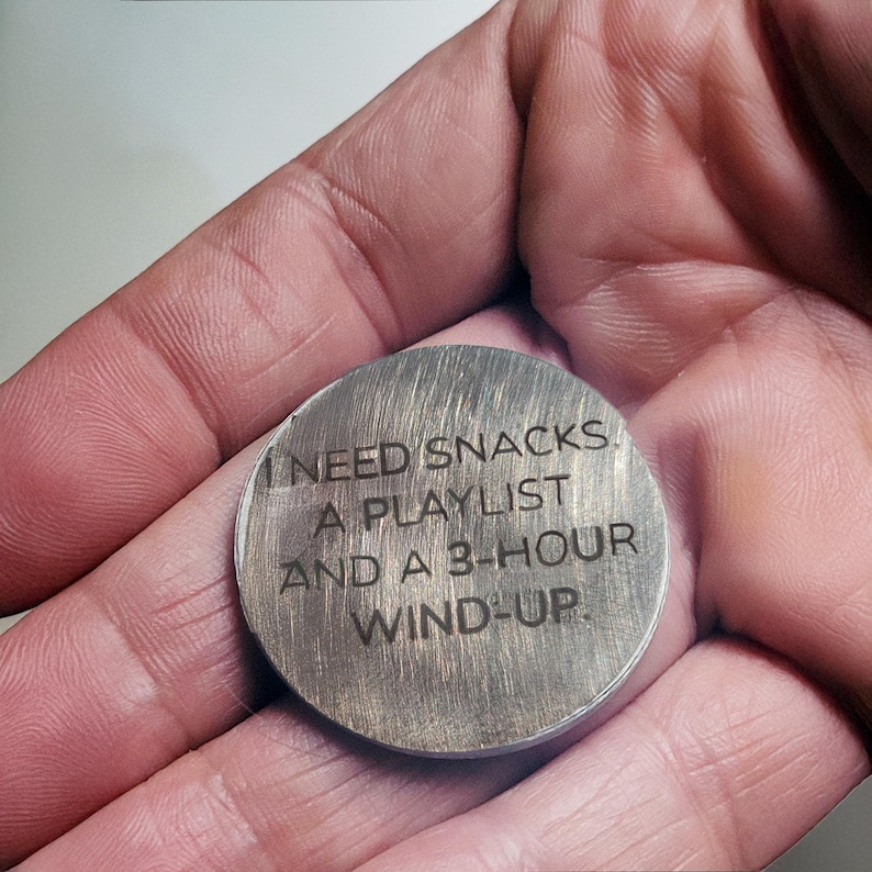 May include: A round, brushed metal token with the engraved text: "I NEED SNACKS, A PLAYLIST AND A 3-HOUR WIND-UP." The token is held in a person's hand, showcasing its size and texture. The overall theme is relaxation and leisure.