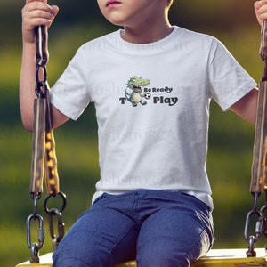 Op de afbeelding: Wit T-shirt met een groene cartoon dinosaurus die een voetbal vasthoudt. De tekst "Be Ready To Play" staat op het shirt gedrukt.