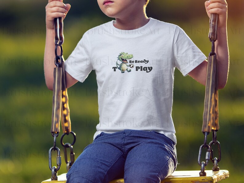 Op de afbeelding: Wit T-shirt met een groene cartoon dinosaurus die een voetbal vasthoudt. De tekst "Be Ready To Play" staat op het shirt gedrukt.