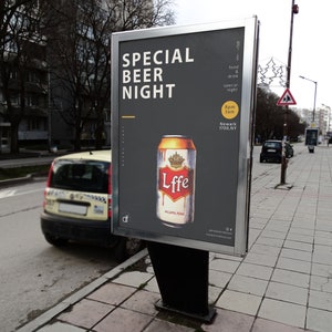 Op de afbeelding: Een zwart en grijs bord met witte tekst die "Special Beer Night" zegt. Het bord toont ook een blikje Lffe-bier met een gouden kroon op het etiket. Het bord staat op een straathoek met een gele auto op de achtergrond.