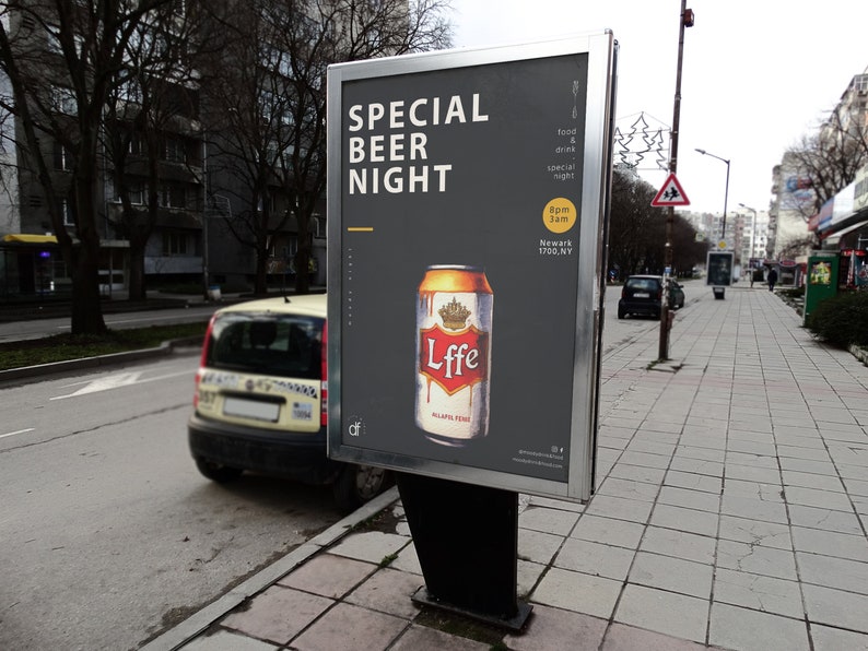 Op de afbeelding: Een zwart en grijs bord met witte tekst die "Special Beer Night" zegt. Het bord toont ook een blikje Lffe-bier met een gouden kroon op het etiket. Het bord staat op een straathoek met een gele auto op de achtergrond.